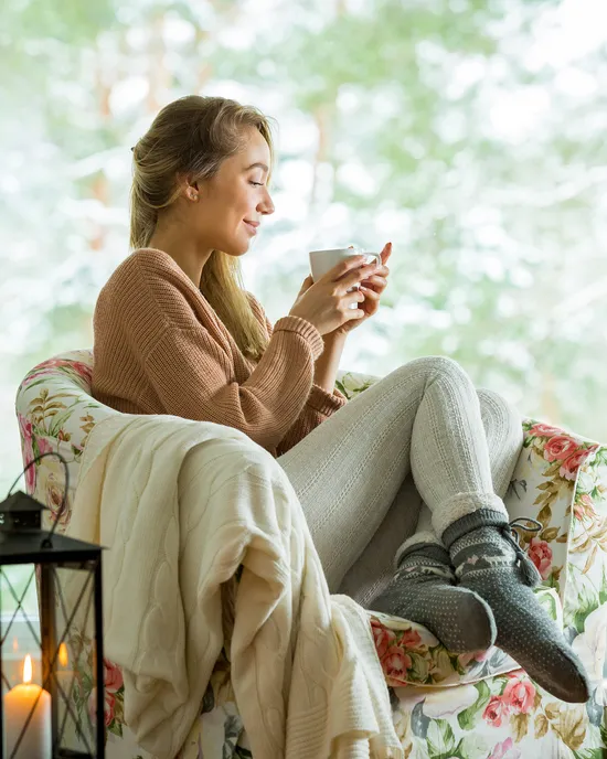 Frau in gemütlichem Sessel trinkt Kaffee am verschneiten Fenster mit Kerzen und Pflanzen