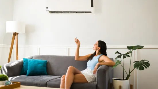 Frau auf grauem Sofa steuert Klimaanlage mit Fernbedienung im modernen Wohnzimmer