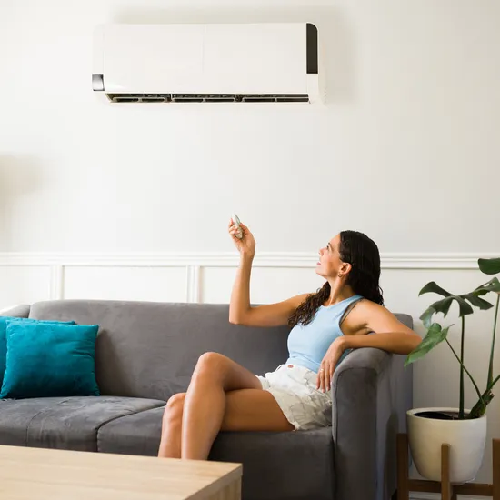 Frau auf grauem Sofa steuert Klimaanlage mit Fernbedienung im modernen Wohnzimmer