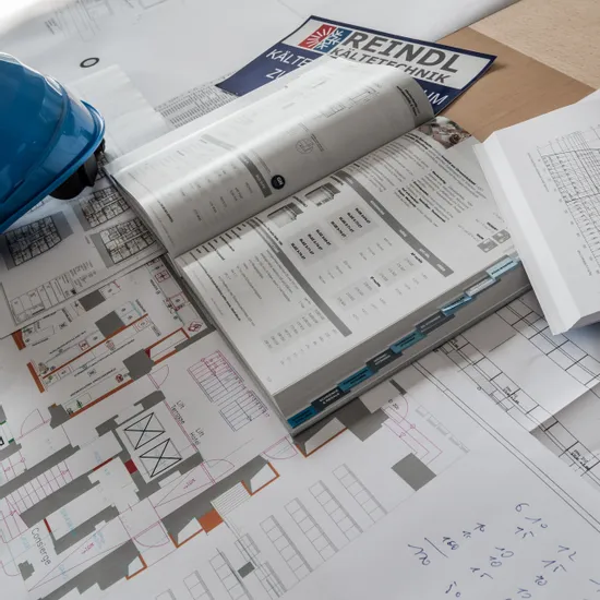 Architektonische Baupläne und offener Katalog auf einem Schreibtisch mit blauem Helm