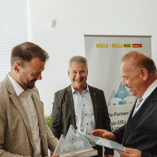 Drei Geschäftsleute teilen Dokumente vor einem Billa Plus Banner