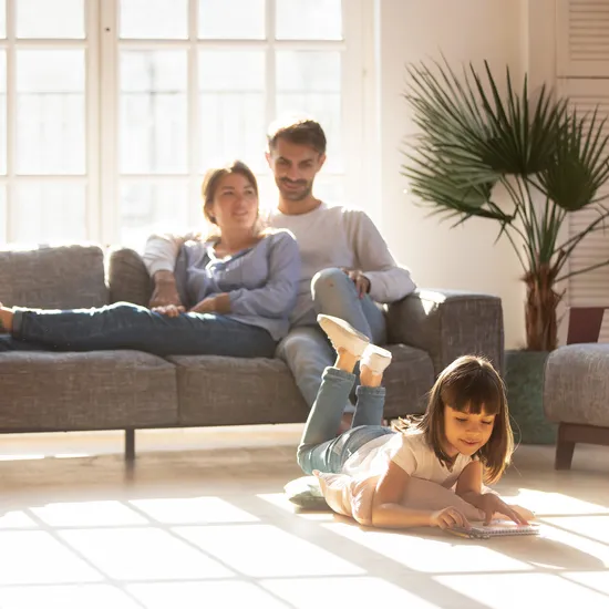 Junges Mädchen zeichnet auf dem Boden, Eltern entspannen auf dem Sofa im sonnigen Wohnzimmer