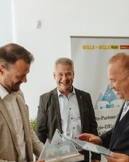 Drei Geschäftsleute teilen Dokumente vor einem Billa Plus Banner