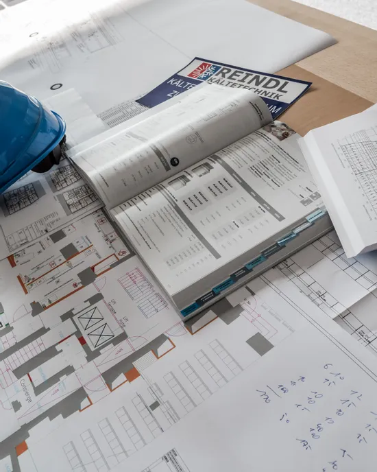 Architektonische Baupläne und offener Katalog auf einem Schreibtisch mit blauem Helm