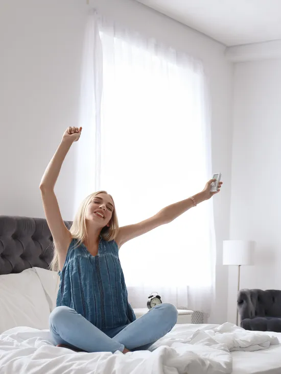 Frau im blauen Oberteil streckt sich freudig auf einem Bett in hellem Raum