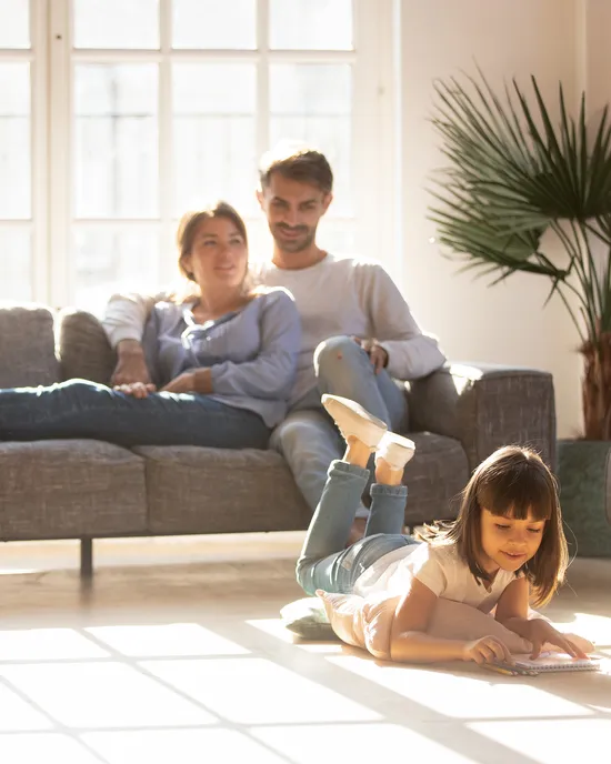 Junges Mädchen zeichnet auf dem Boden, Eltern entspannen auf dem Sofa im sonnigen Wohnzimmer